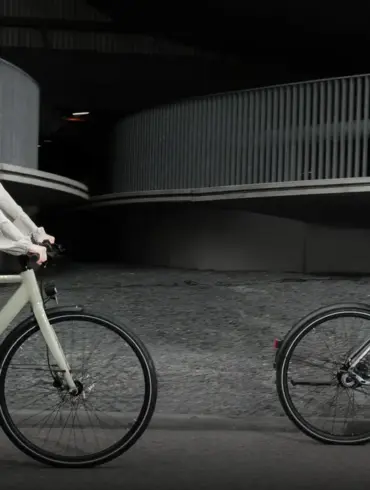 Mann und Frau fahren Tenways CGO600 E-Bikes nebeneinander auf einer Straße, mit moderner Architektur im Hintergrund.