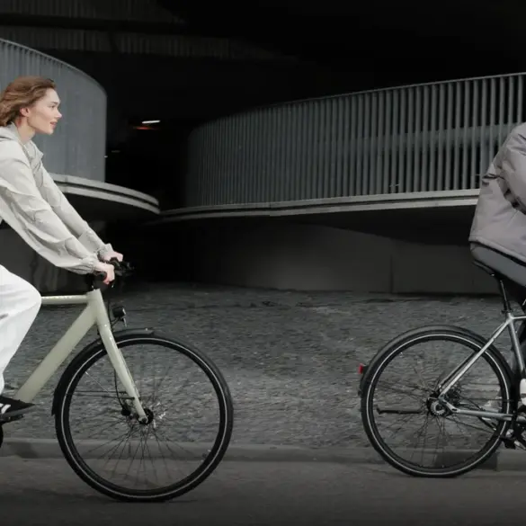 Mann und Frau fahren Tenways CGO600 E-Bikes nebeneinander auf einer Straße, mit moderner Architektur im Hintergrund.