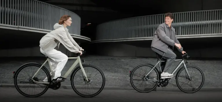 Mann und Frau fahren Tenways CGO600 E-Bikes nebeneinander auf einer Straße, mit moderner Architektur im Hintergrund.
