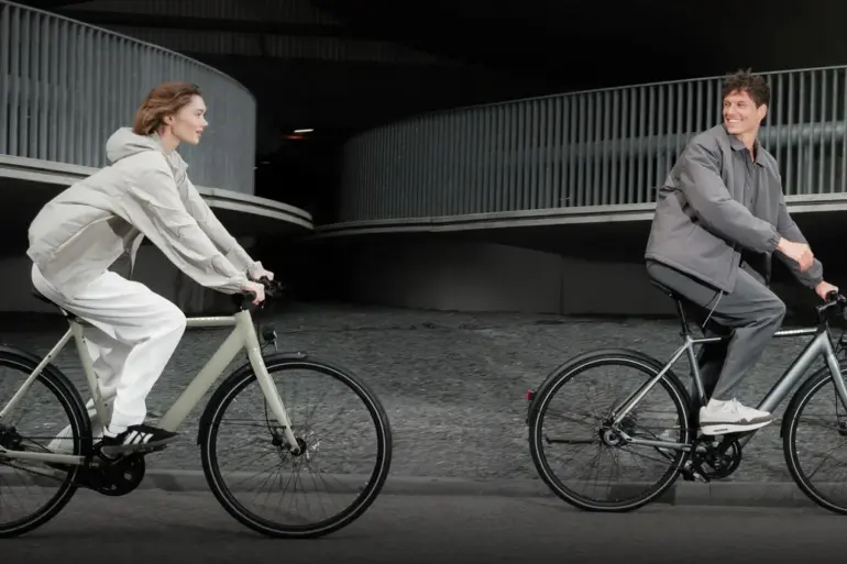 Mann und Frau fahren Tenways CGO600 E-Bikes nebeneinander auf einer Straße, mit moderner Architektur im Hintergrund.