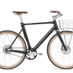 Schwarz mattes Arthur Cruiser E-Bike mit braunem Ledersattel, braunen Griffen und einem silbernen Frontkorb, ausgestattet mit Schutzblechen und einem Zahnriemenantrieb.