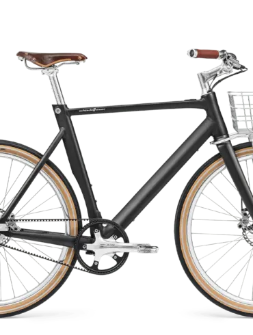 Schwarz mattes Arthur Cruiser E-Bike mit braunem Ledersattel, braunen Griffen und einem silbernen Frontkorb, ausgestattet mit Schutzblechen und einem Zahnriemenantrieb.