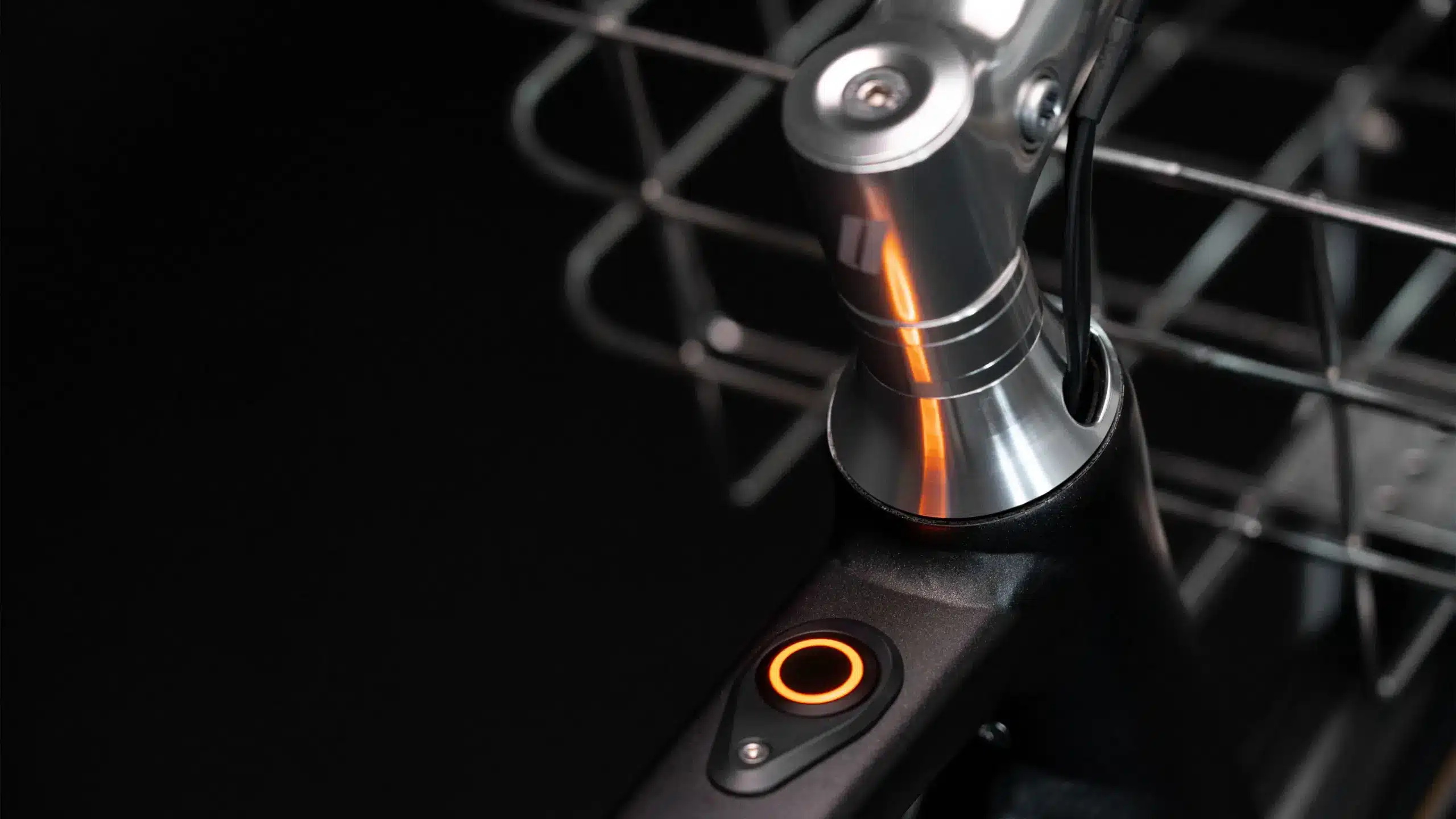 Nahaufnahme des Arthur Cruisers E-Bikes, Fokus auf den beleuchteten orangefarbenen runden Knopf am Rahmen und Teile des Lenkers sowie eines Einkaufskorbs im Hintergrund.