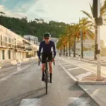 Ein Mann fährt mit einem Bergamont E-Grandurance Elite Fahrrad auf einer Küstenstraße, umgeben von Palmen und Gebäuden.