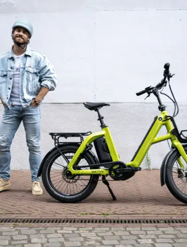 Limegrünes BESV CPT 1.0 E-Bike mit vorne montiertem Gepäckträger und hinterem Gepäckträger, daneben ein Mann mit Jeansjacke und Helm.