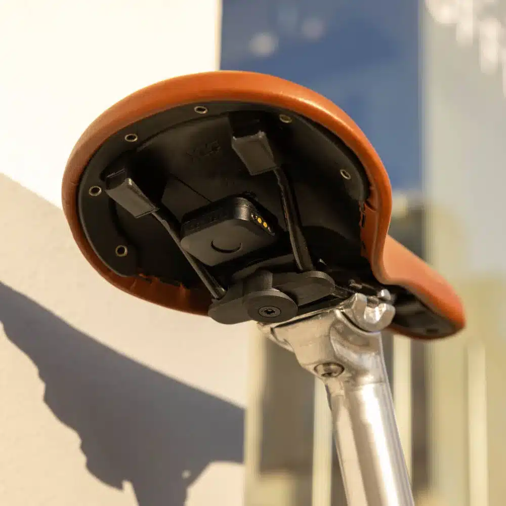 Detailaufnahme der Unterseite eines braunen Leder-Fahrradsattels, an dem das schwarze Quad Lock Alarm & Finder-Gerät befestigt ist. Das Gerät ist mit Kabeln verbunden und die Halterung ist sichtbar.