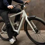Mann fährt ein hellgraues Amsterdam Urban E-Bike mit schwarzer Kleidung und Sneakers auf einem rustikalen Hintergrund.