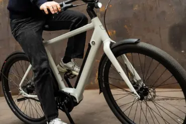 Mann fährt ein hellgraues Amsterdam Urban E-Bike mit schwarzer Kleidung und Sneakers auf einem rustikalen Hintergrund.
