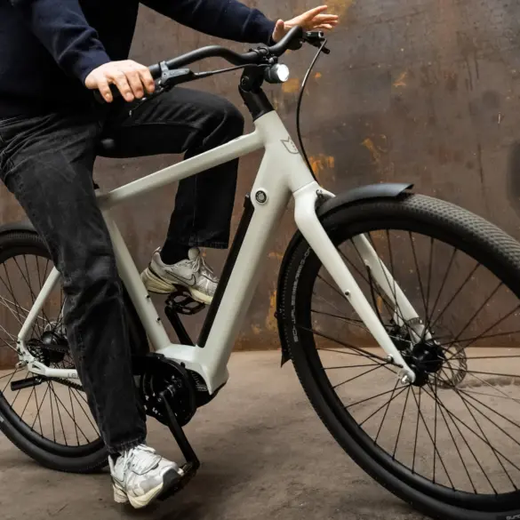 Mann fährt ein hellgraues Amsterdam Urban E-Bike mit schwarzer Kleidung und Sneakers auf einem rustikalen Hintergrund.