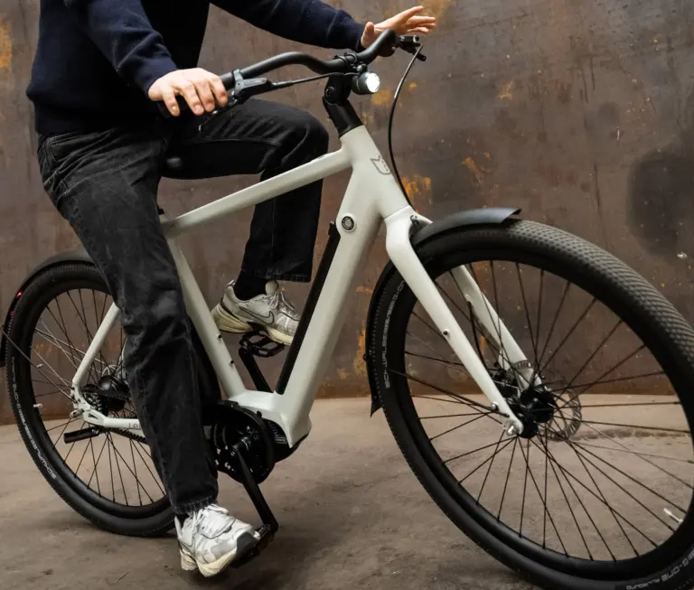 Mann fährt ein hellgraues Amsterdam Urban E-Bike mit schwarzer Kleidung und Sneakers auf einem rustikalen Hintergrund.