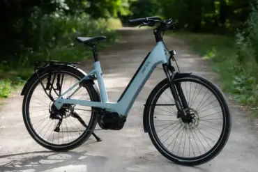 Ein hellblaues Antwerp PX 9 City-E-Bike mit Gepäckträger und breiten Reifen steht auf einem Feldweg, umgeben von grünen Bäumen.