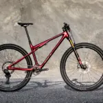Seitenansicht des roten BH LYNX RACE 8.0 Mountainbikes mit Vollfederung und Offroad-Reifen, stehend auf Asphalt vor einer felsigen Kulisse.