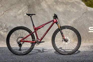 Seitenansicht des roten BH LYNX RACE 8.0 Mountainbikes mit Vollfederung und Offroad-Reifen, stehend auf Asphalt vor einer felsigen Kulisse.