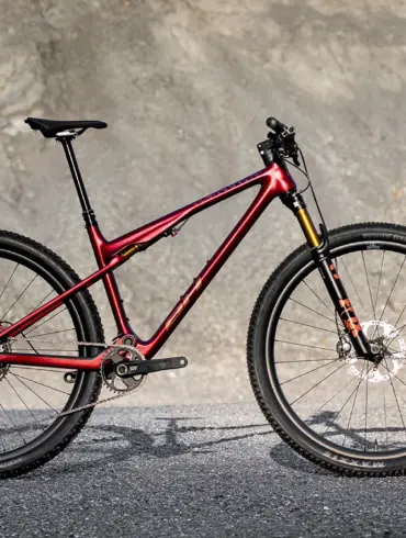 Seitenansicht des roten BH LYNX RACE 8.0 Mountainbikes mit Vollfederung und Offroad-Reifen, stehend auf Asphalt vor einer felsigen Kulisse.