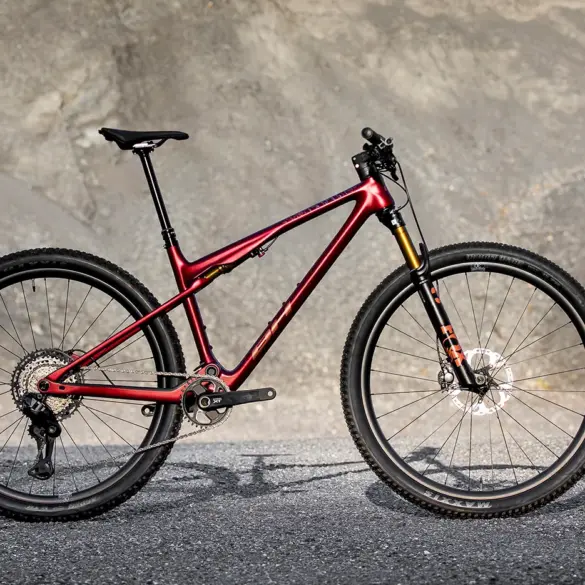 Seitenansicht des roten BH LYNX RACE 8.0 Mountainbikes mit Vollfederung und Offroad-Reifen, stehend auf Asphalt vor einer felsigen Kulisse.