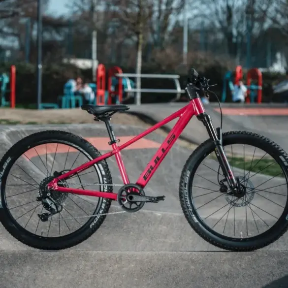 Seitenansicht eines pinkfarbenen BULLS Tokee Disc 24 Kinderfahrrads mit Scheibenbremsen, das auf einer Betonrampe vor einem Spielplatz steht.