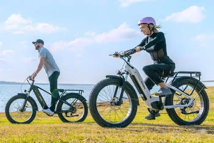 Mann auf schwarzem HAOQI Eagle E-Bike, Frau auf weißem HAOQI Eagle E-Bike, beide fahren auf einem grasbewachsenen Ufer in der Nähe eines Gewässers unter bewölktem Himmel.