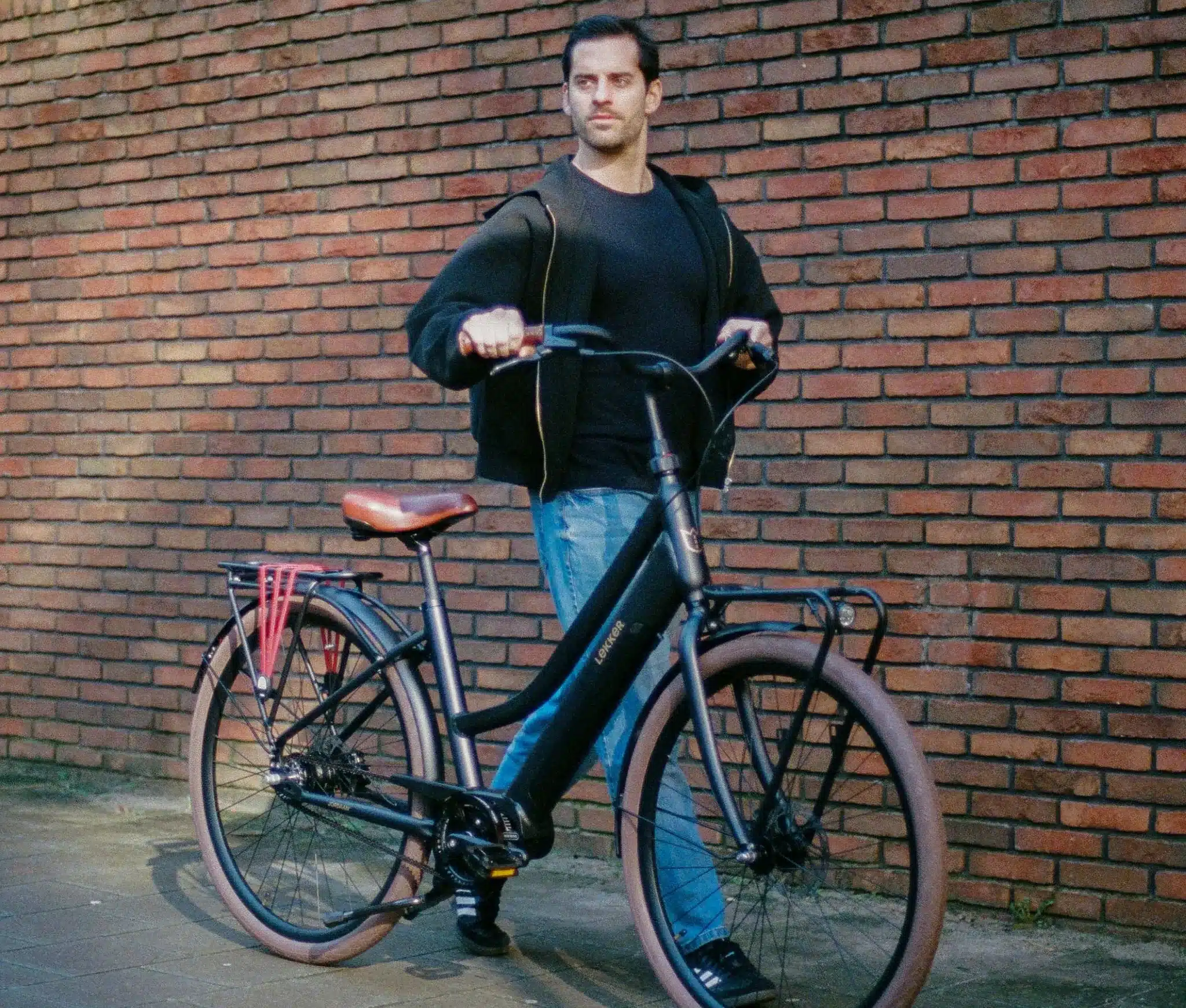 Ein Mann steht neben einem schwarzen LEKKER Jordaan Urban E-Bike mit braunem Sattel und Kofferträgern.