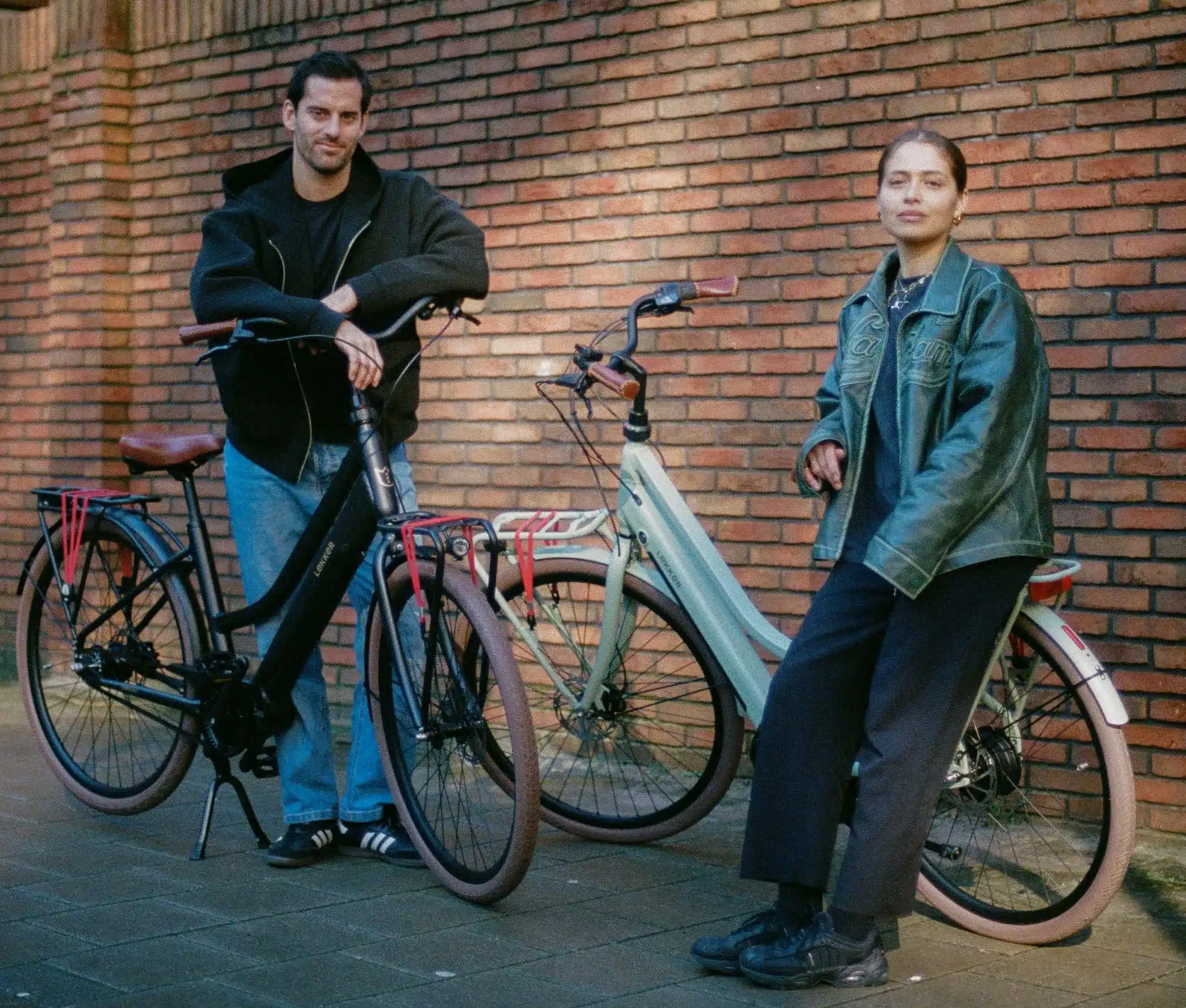Mann und Frau stehen neben zwei LEKKER Jordaan Urban E-Bikes vor einer Ziegelmauer.