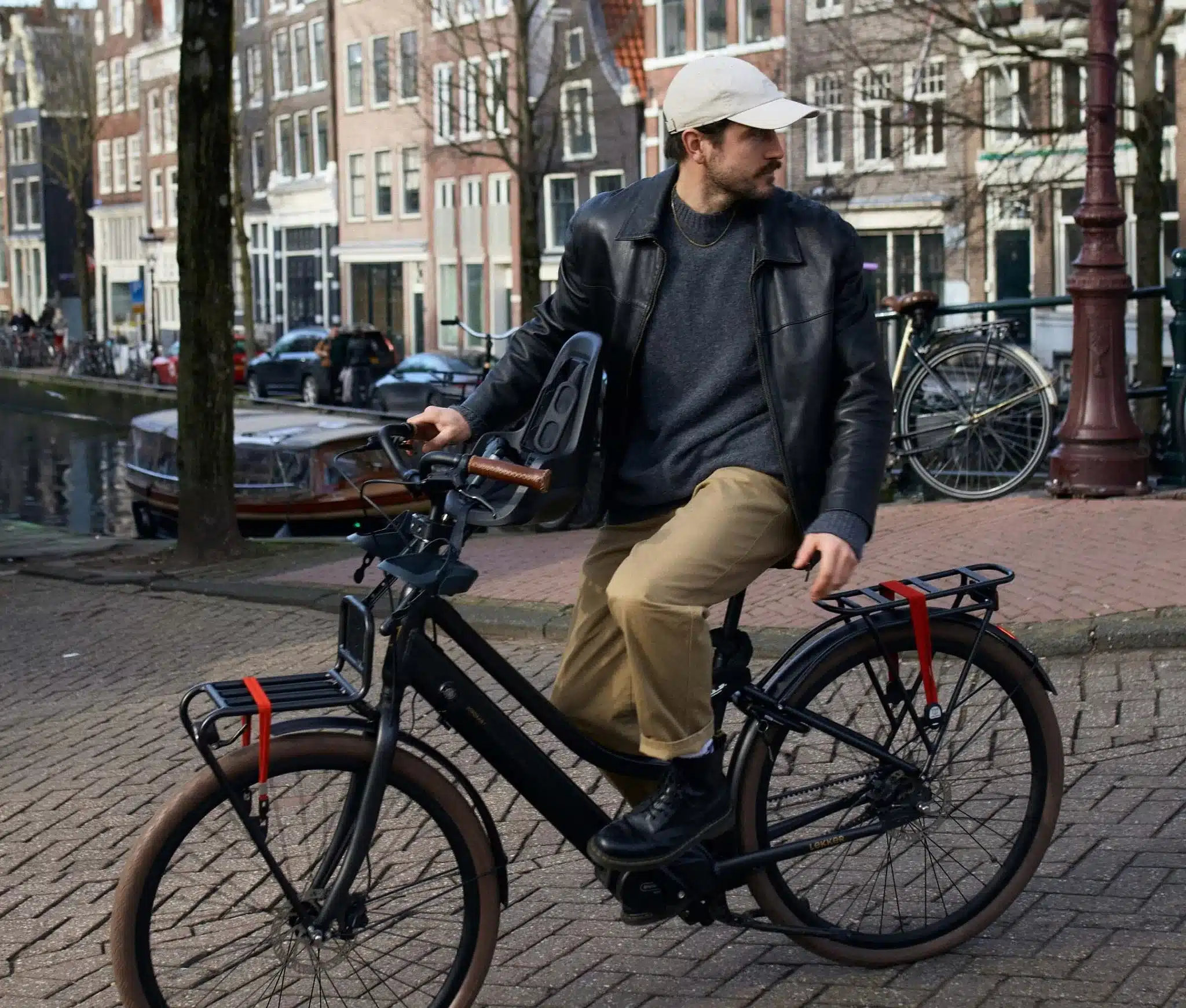 Ein Mann mit Kappe und Lederjacke fährt ein schwarzes LEKKER Jordaan Urban eBike mit Kindersitz über ein Kopfsteinpflaster in einer Stadt mit Kanälen im Hintergrund.