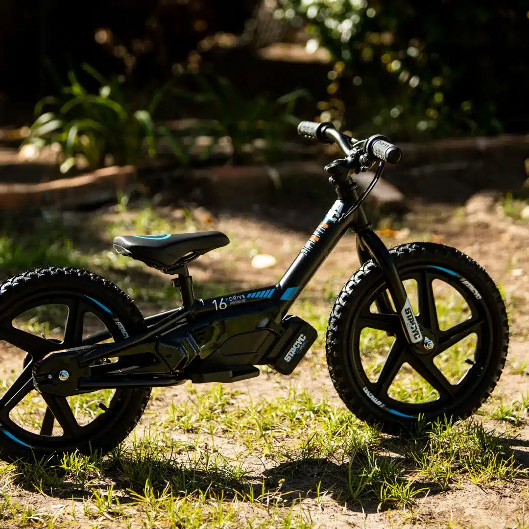 Ein schwarzes STACYC 16eDRIVE BRUSHLESS Kinderfahrrad mit blauen Akzenten steht auf Gras im Freien. Das Fahrrad ist für Kinder gedacht, die vom Laufrad zum motorisierten Fahren wechseln.