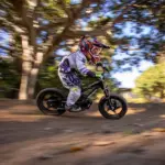 Ein Kind mit Helm und Schutzkleidung fährt auf einem schwarzen STACYC 16eDRIVE ELITE Elektro-Balancebike auf einem Feldweg unter Bäumen.