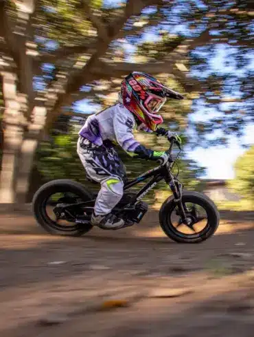 Ein Kind mit Helm und Schutzkleidung fährt auf einem schwarzen STACYC 16eDRIVE ELITE Elektro-Balancebike auf einem Feldweg unter Bäumen.