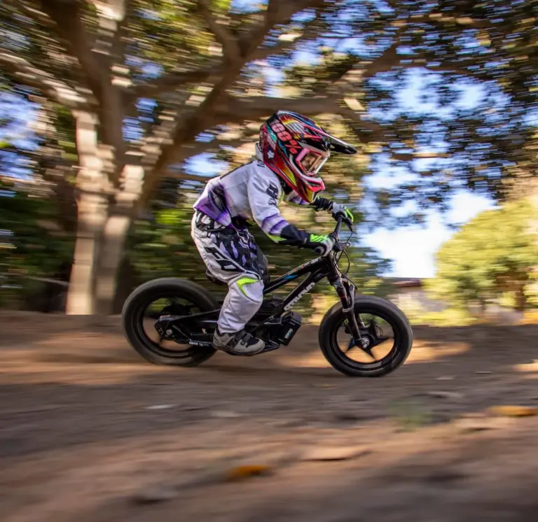Ein Kind mit Helm und Schutzkleidung fährt auf einem schwarzen STACYC 16eDRIVE ELITE Elektro-Balancebike auf einem Feldweg unter Bäumen.