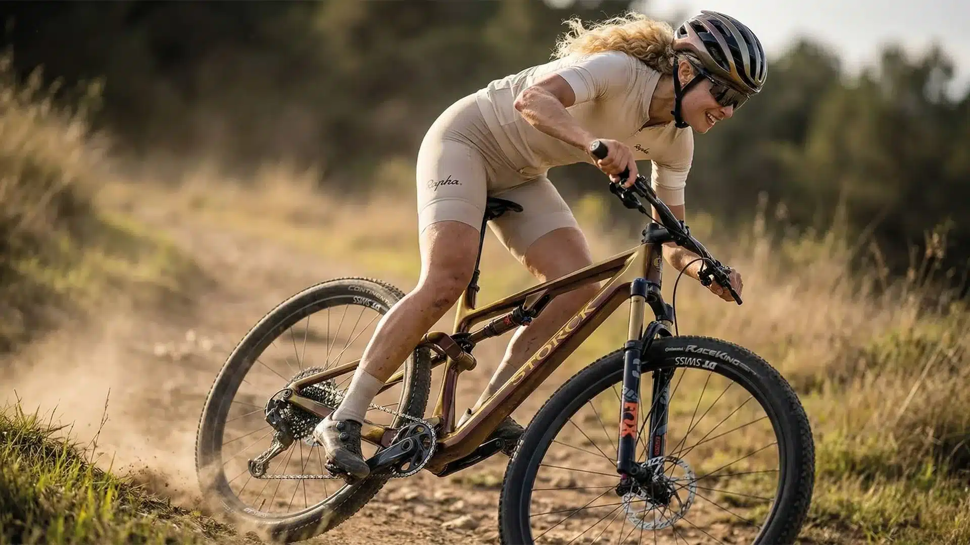 Eine Frau mit Helm und Sportbekleidung fährt ein braunes Storck Adrenalin Mountainbike auf einem Feldweg, Staub wirbelt auf.