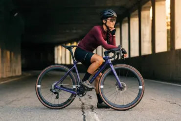 Frau mit Fahrrad, Storck Grix.3 Gravelbike, lila Rahmen, vor einer dunklen Betonwand stehend