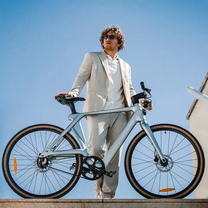 Ein Mann in einem hellen Anzug präsentiert das schlanke Fiido Air E-Bike vor einem blauen Himmel.