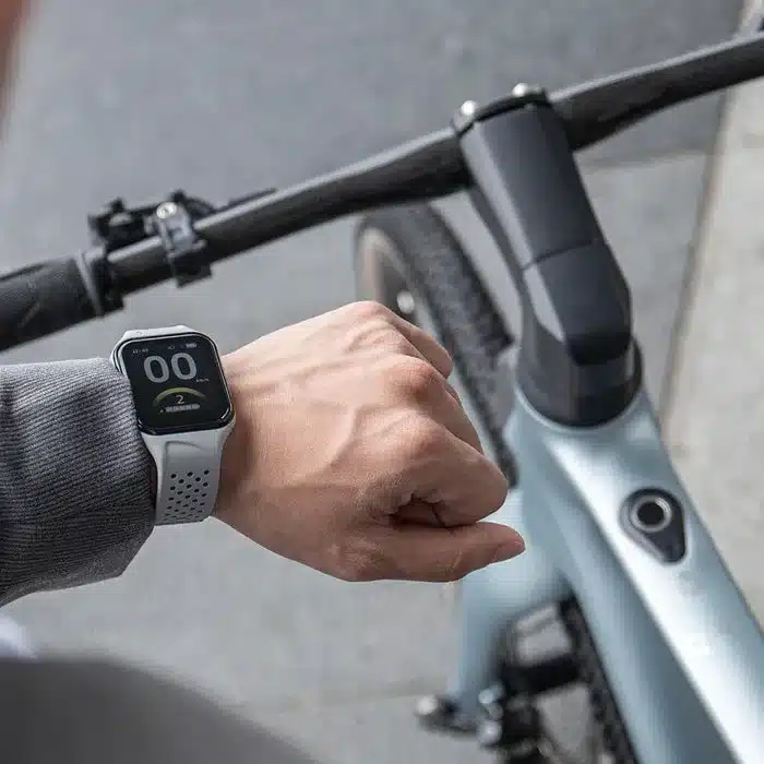 Nahaufnahme der Hand eines Mannes, der eine Smartwatch trägt, die Informationen über das Fiido Air E-Bike anzeigt. Das E-Bike ist im Hintergrund zu sehen.