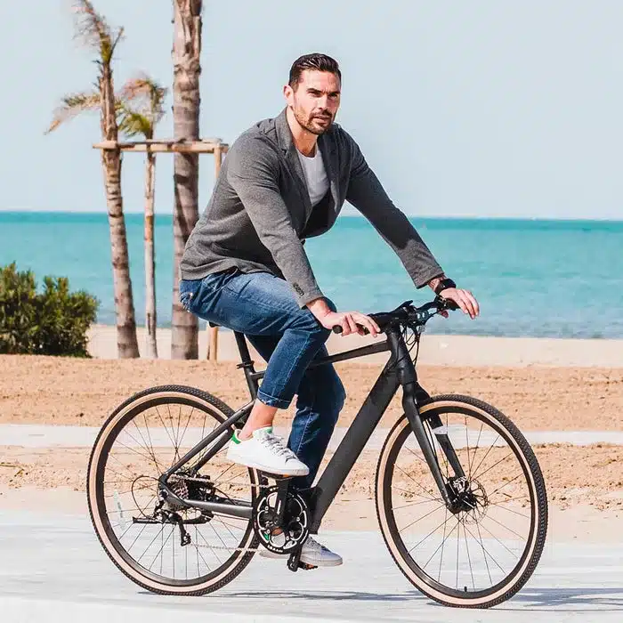 Ein Mann fährt ein schwarzes Fiido C21 E-Bike auf einer Promenade am Meer, mit Sandstrand und Palmen.