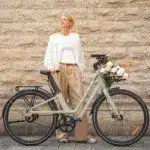 Eine Frau posiert lächelnd neben einem hellgrünen Fiido C700 City E-Bike, das vorne mit einem Korb voller Rosen geschmückt ist. Das Fahrrad hat einen Riemenantrieb und eine klassische Optik.