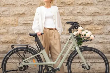 Eine Frau posiert lächelnd neben einem hellgrünen Fiido C700 City E-Bike, das vorne mit einem Korb voller Rosen geschmückt ist. Das Fahrrad hat einen Riemenantrieb und eine klassische Optik.