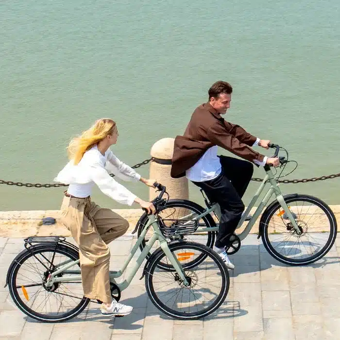 Mann und Frau fahren nebeneinander auf mintgrünen Fiido C700 E-Bikes, die Promenade und das Wasser im Hintergrund.