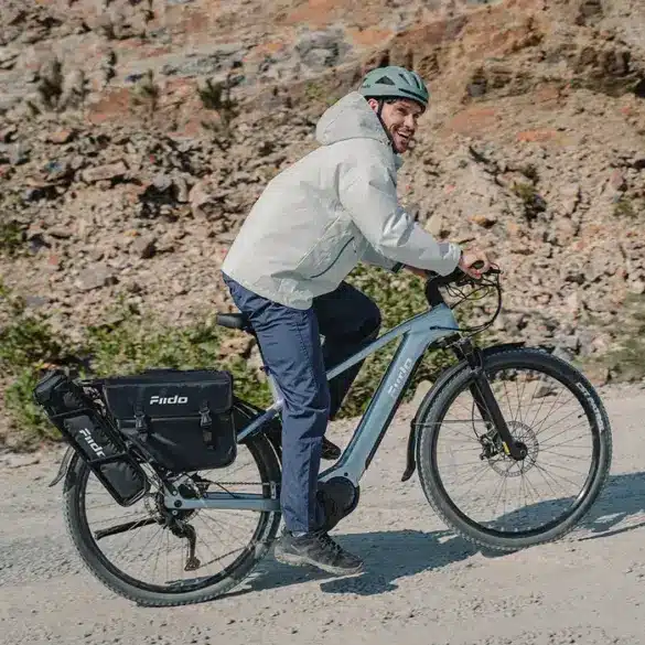 Mann mit Helm fährt Fiido Nomads Pro E-Bike mit Gepäcktaschen auf einem Schotterweg bergauf.