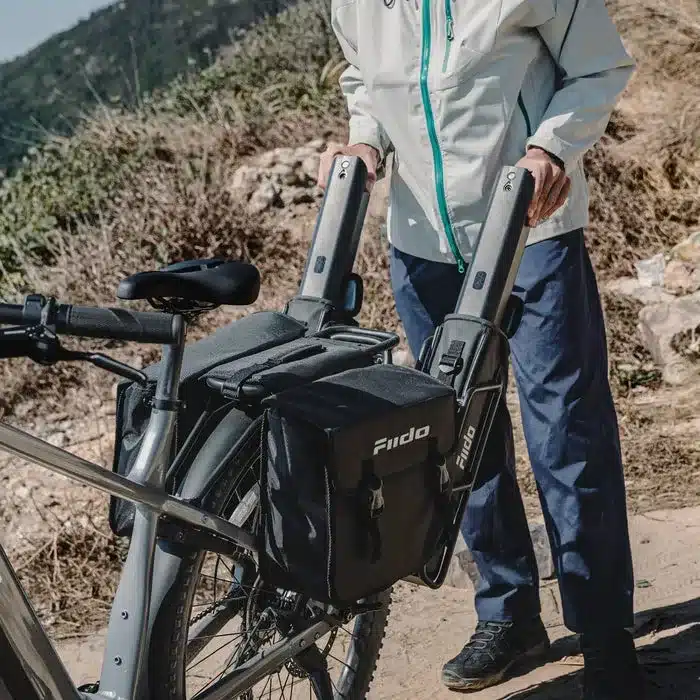 Eine Person lädt zwei lange, rechteckige Akkus für ein Fiido Nomads Pro E-Bike. Die Akkus sind in schwarzen Halterungen an der Seite des Fahrrads befestigt. Das Fahrrad ist mit einer Federgabel und Packtaschen ausgestattet.