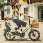 Eine Frau fährt mit einem Kind auf einem grünen Fiido T3 Lastenrad. Das Kind sitzt sicher in einem Kindersitz auf dem Gepäckträger und trägt einen grauen Helm. Vorne am Lenker ist ein Weidenkorb mit Brot und Äpfeln befestigt.