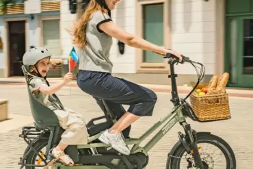 Eine Frau fährt mit einem Kind auf einem grünen Fiido T3 Lastenrad. Das Kind sitzt sicher in einem Kindersitz auf dem Gepäckträger und trägt einen grauen Helm. Vorne am Lenker ist ein Weidenkorb mit Brot und Äpfeln befestigt.