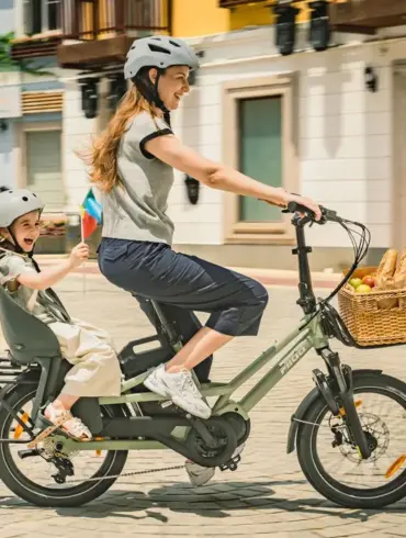 Eine Frau fährt mit einem Kind auf einem grünen Fiido T3 Lastenrad. Das Kind sitzt sicher in einem Kindersitz auf dem Gepäckträger und trägt einen grauen Helm. Vorne am Lenker ist ein Weidenkorb mit Brot und Äpfeln befestigt.