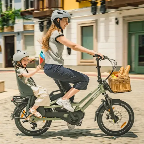 Eine Frau fährt mit einem Kind auf einem grünen Fiido T3 Lastenrad. Das Kind sitzt sicher in einem Kindersitz auf dem Gepäckträger und trägt einen grauen Helm. Vorne am Lenker ist ein Weidenkorb mit Brot und Äpfeln befestigt.