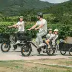 Ein Mann fährt ein beiges Fiido T3 Max Longtail-Fahrrad mit zwei Kindern auf Kindersitzen im hinteren Bereich und zieht einen schwarzen Anhänger hinter sich her. Eine Frau fährt ein ähnliches Fahrrad daneben. Beide Fahrräder sind auf einer asphaltierten Straße in einer landschaftlich reizvollen Gegend unterwegs.