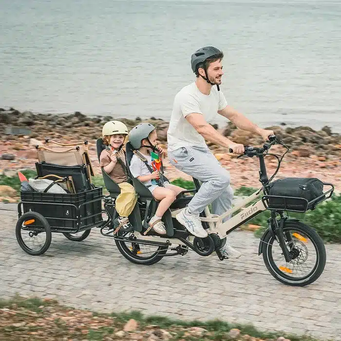 Vater fährt ein Fiido T3 Max Longtail-E-Bike mit zwei Kindern in Kindersitzen und einem Anhänger am Meer.