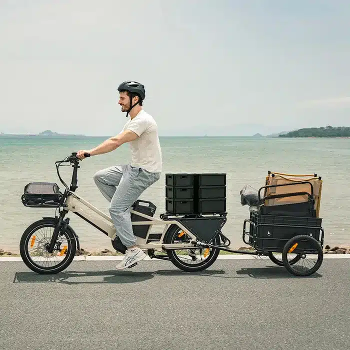 Mann fährt Fiido T3 Max Longtail E-Bike mit Anhänger, beladen mit Kisten und Ausrüstung am Meer.