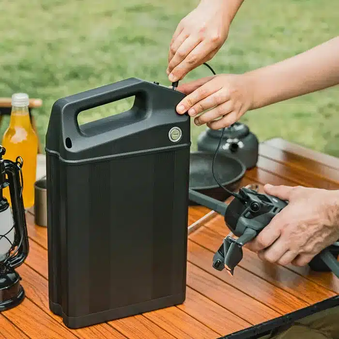 Eine Person schließt ein Kabel an einen Anschluss an der Oberseite eines großen, schwarzen Batterie-Packs an, das auf einem Holztisch steht.