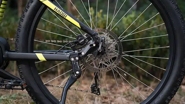 Detailaufnahme des Hinterrads eines HAOQI Cheetah E-MTB. Man sieht die Nabe, die Speichen, die Bremsscheibe und den Schaltwerk mit Kette. Am schwarzen Rahmen sind gelbe Streifen zu erkennen.