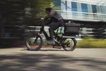 Fahrer eines schwarzen Lemmo 3 Longtail-E-Bikes mit einem Kind oder Haustier in einer Gepäckbox fährt auf einer Straße.