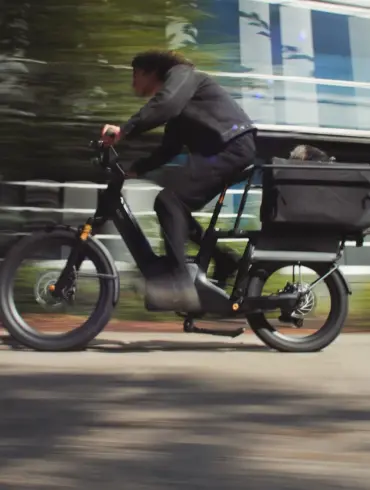 Fahrer eines schwarzen Lemmo 3 Longtail-E-Bikes mit einem Kind oder Haustier in einer Gepäckbox fährt auf einer Straße.