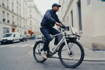 Ein Mann fährt ein silbernes Lemmo One "Collab. Ride" E-Bike mit einer Stofftasche über dem Vorderrad und einem Gepäckträger hinten durch eine Stadtstraße.